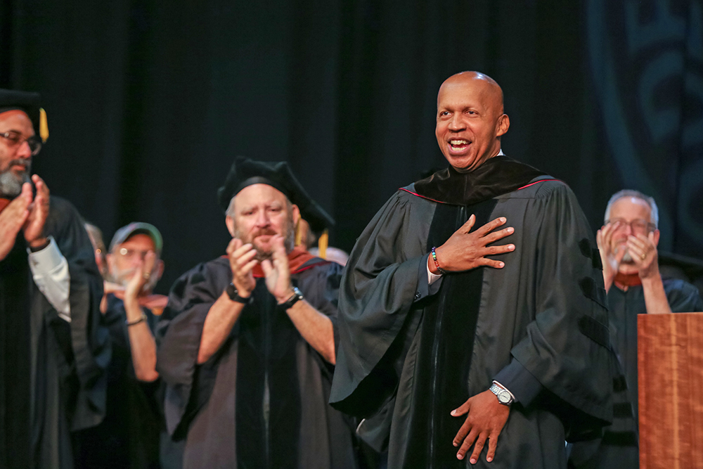 Faculty applaud fellow member on graduation stage.