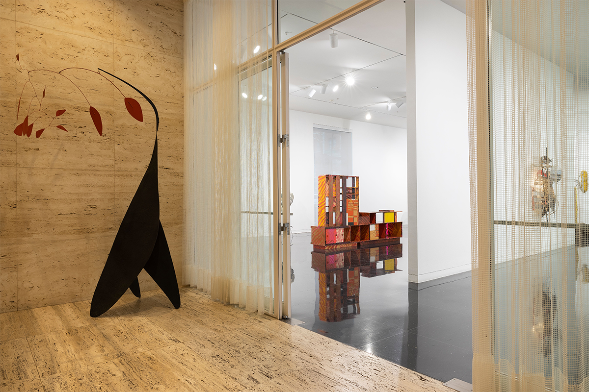 A wood paneled exhibition hall with a three-legged black sculpture and a sculpture made of boxes