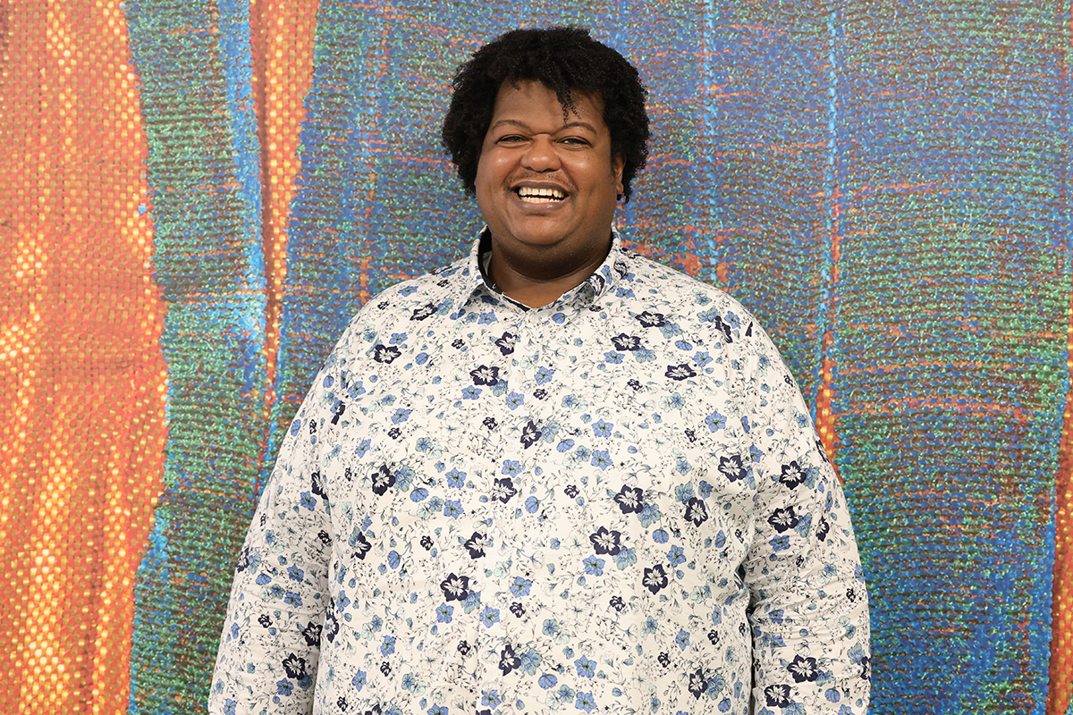 Dieng Cameron stands smiling against a weaving-inspired wallpaper