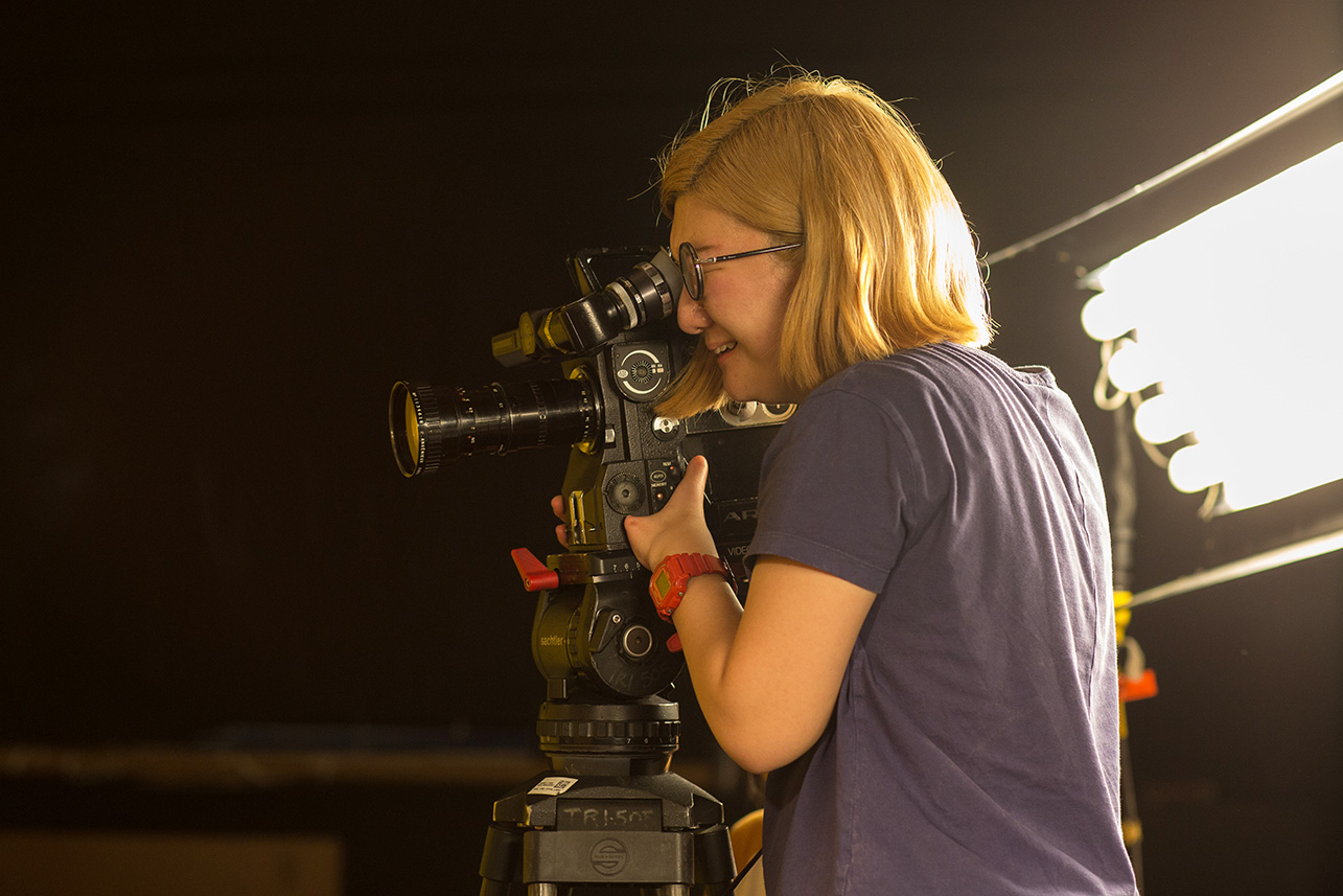 Photo of a woman holding a video camera with a light behind her.