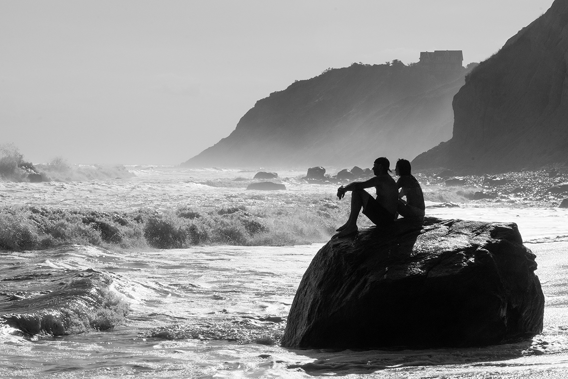 Black and White photo of two people sitting on a rock along the sea shore.