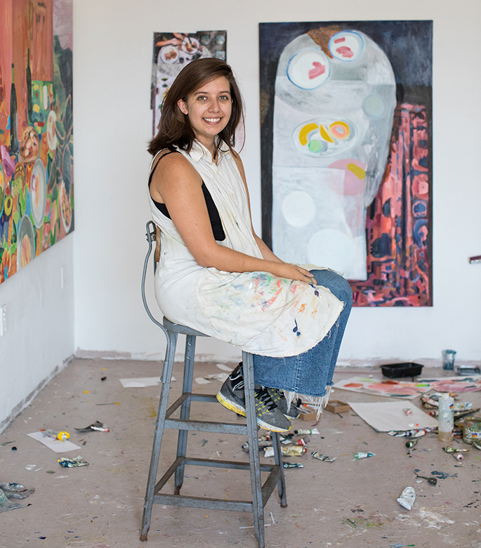 Photo of Kiernan Pazdar seated on a stool with her paintings behind her.