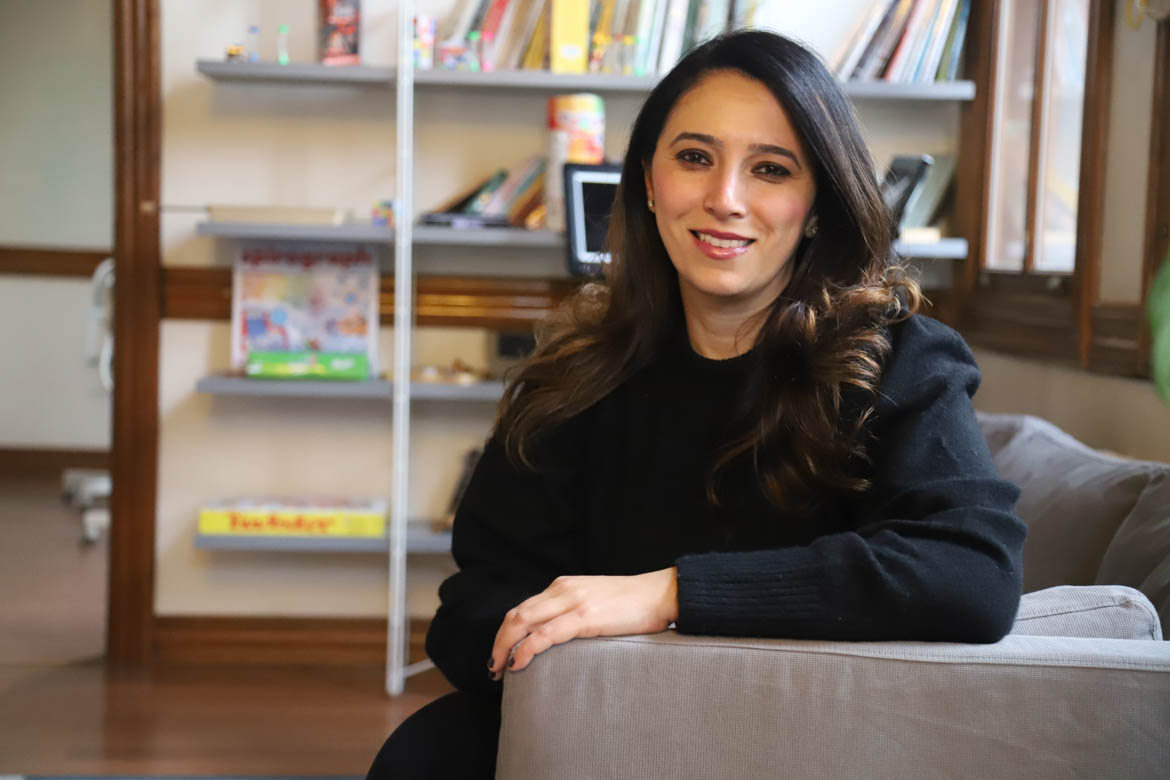 Lina Sergie Attar sits on a couch with a bookshelf behind.