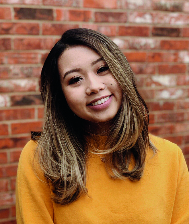 An image of Denni Zhao smiling in front of a red brick background