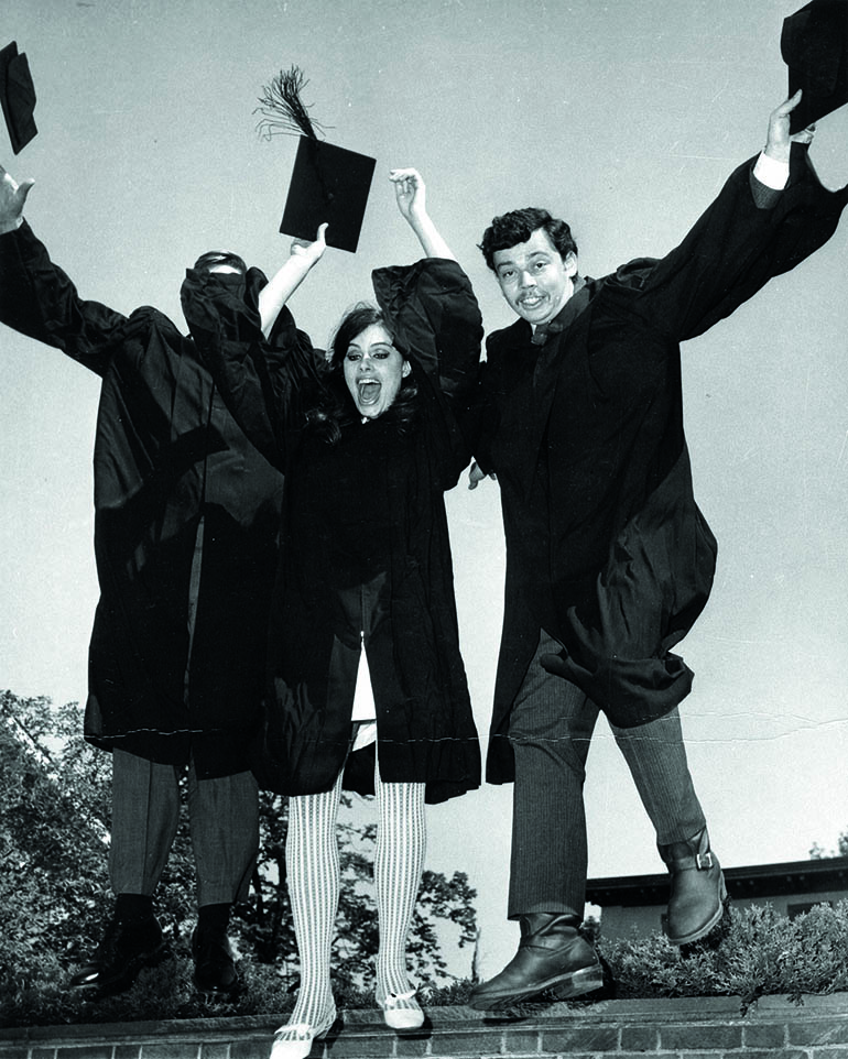 A black and white image of RISD alumni dressed in graduation regalia jumping with their arms in the air and smiling