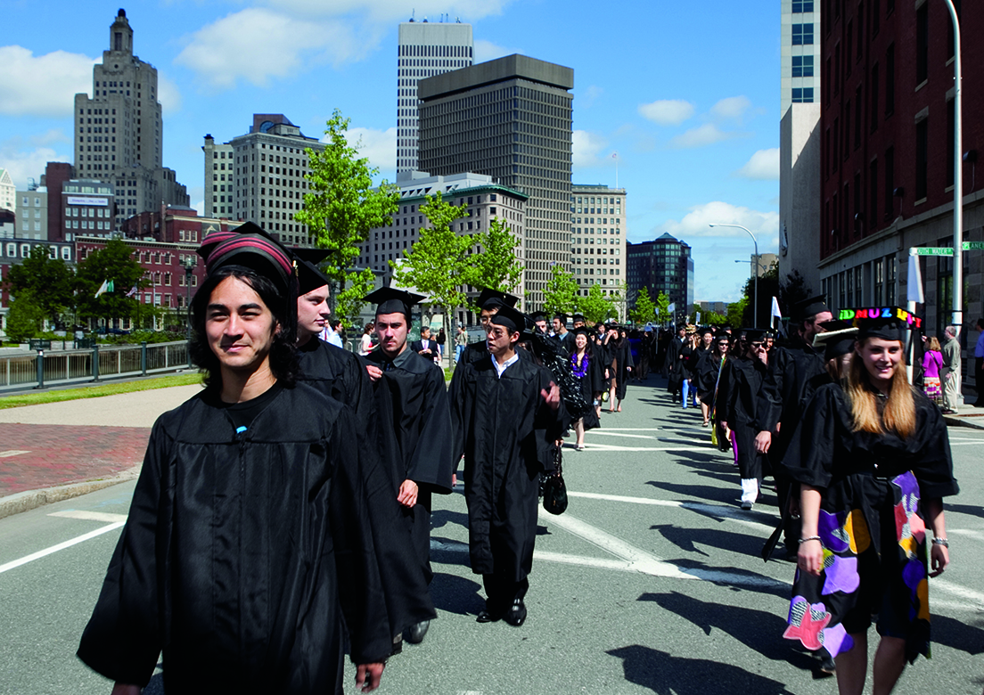 An image of students walking down a sidewalk dressed in graduation regalia