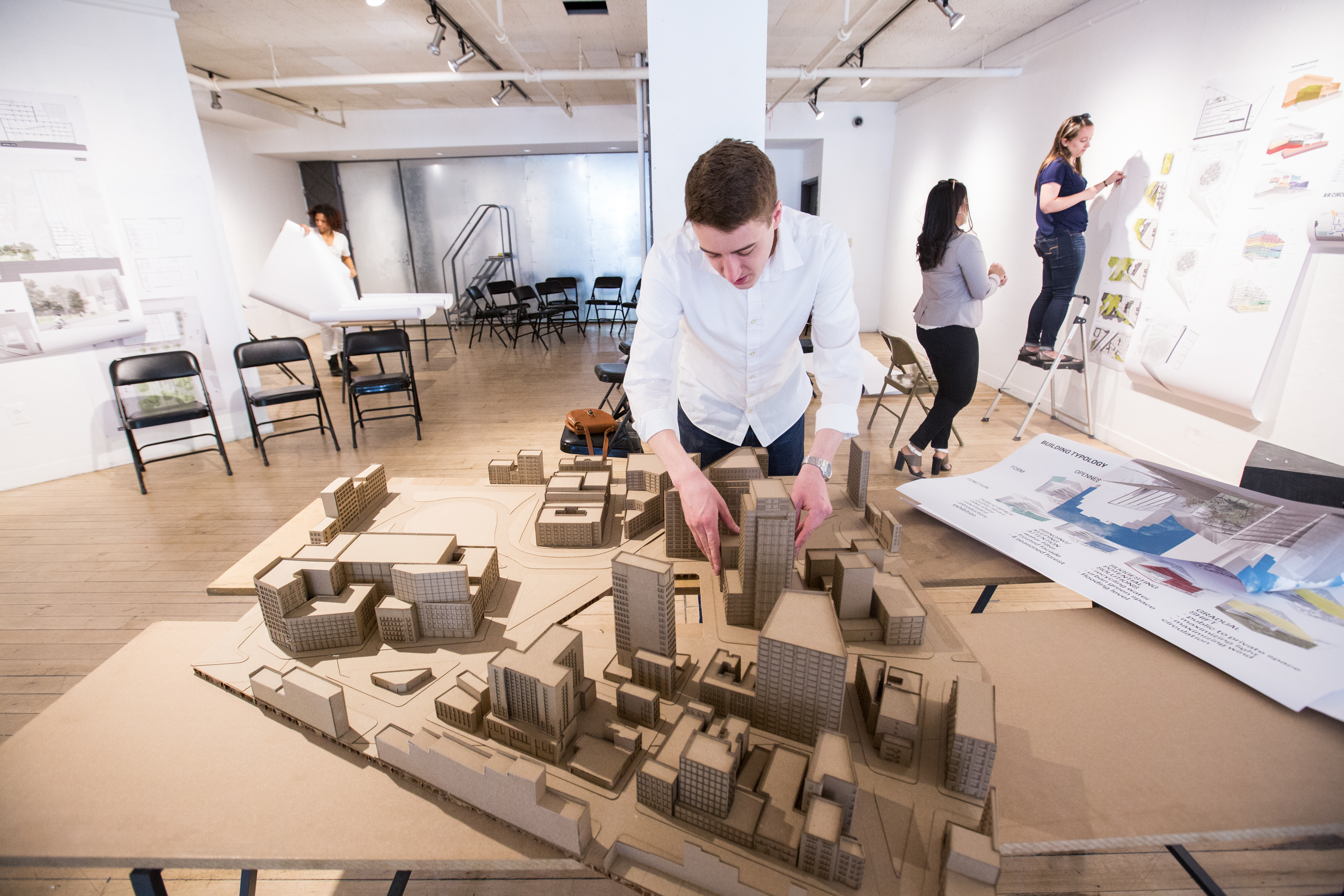 A RISD architecture student working on a cardboard model of an urban scene in a white-walled classroom.