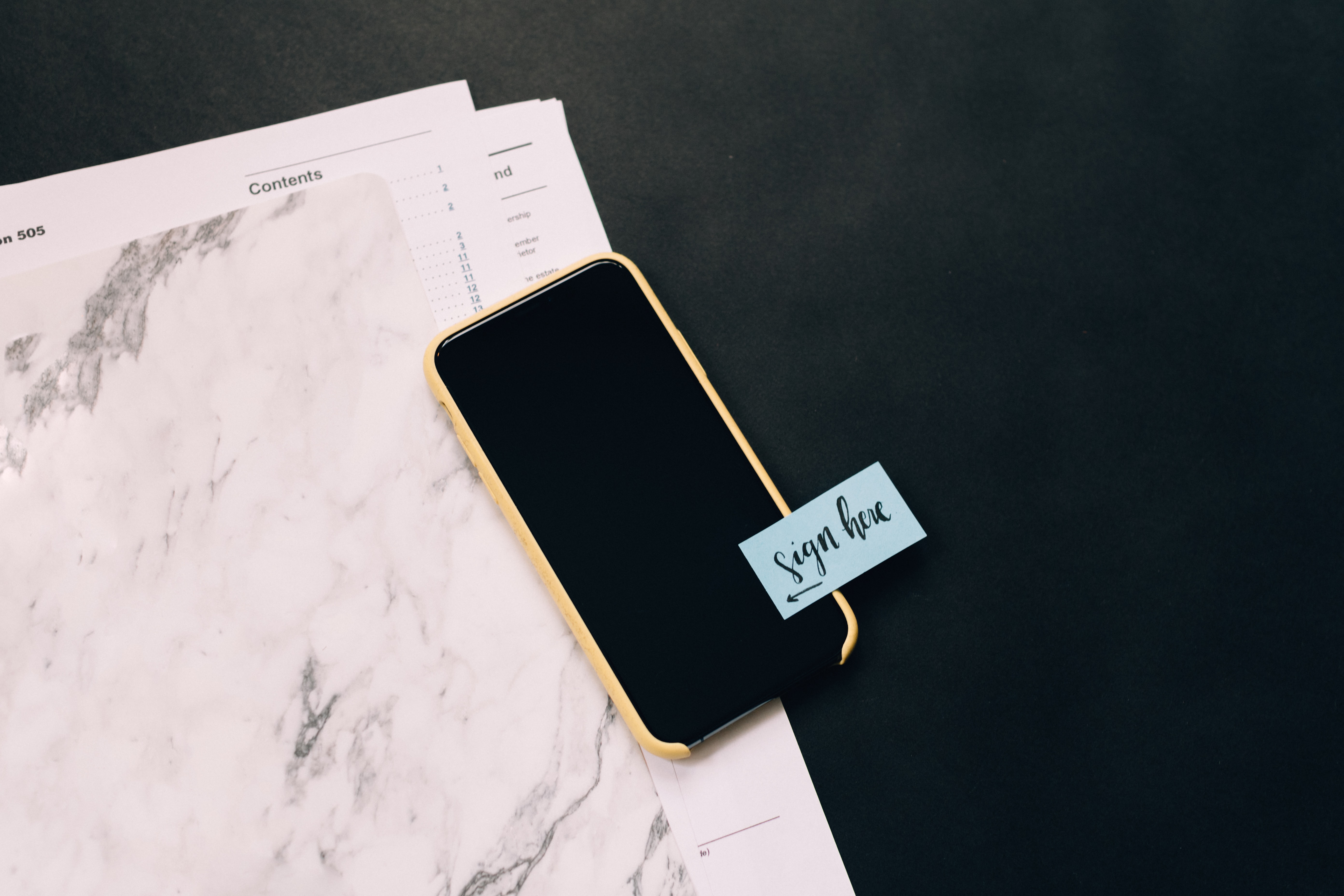 A photo of a smartphone with a "sign here" post-it note attached, laying on top of a folder with government forms in it.
