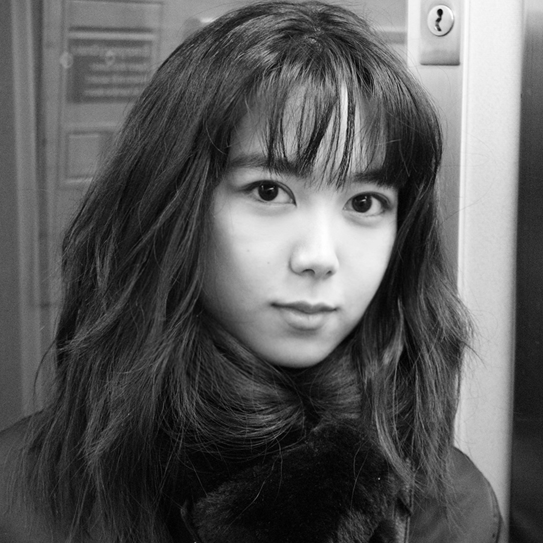 Black + white image of a female with long dark hair and bangs standing in front of school lockers
