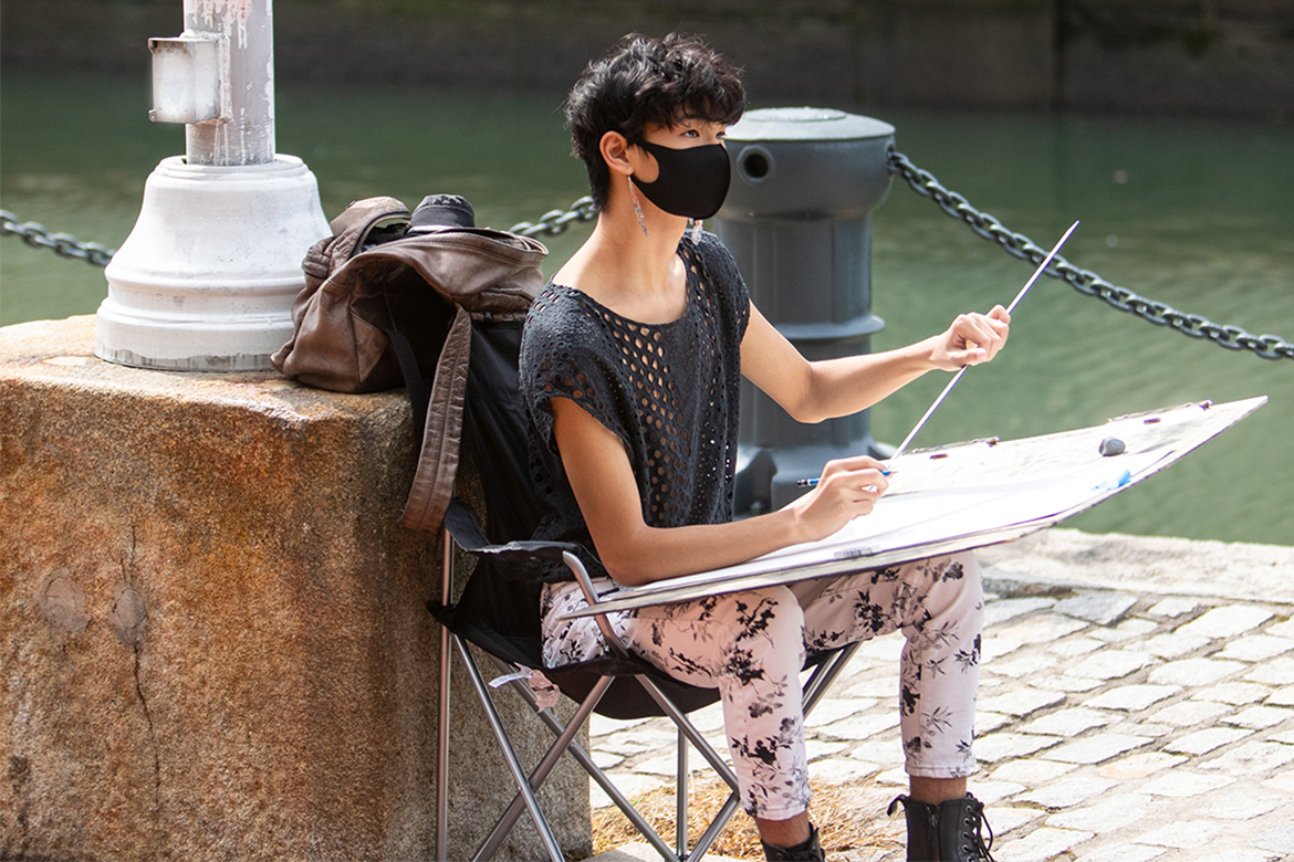 Image of a person in a black mask wearing pink and black leggings, a black shirt, sitting by a river with a drawing board