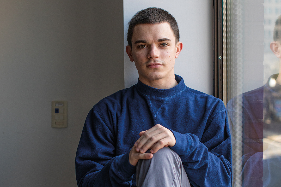 Image of a male sitting in a window with a blue sweater and grey pants