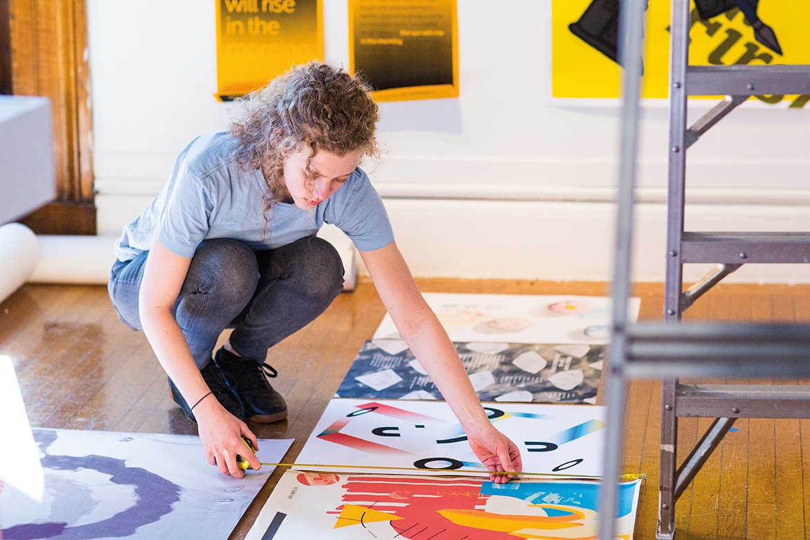 Image of a female in a blue shirt and jeans crouched over paintings laid out on the floor
