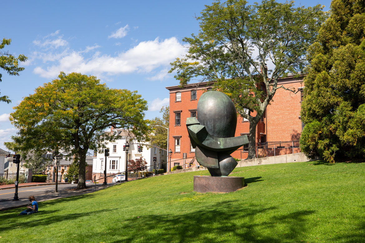 Image of an abstract sculpture on a green lawn in front of orange brick building with trees and blue sky