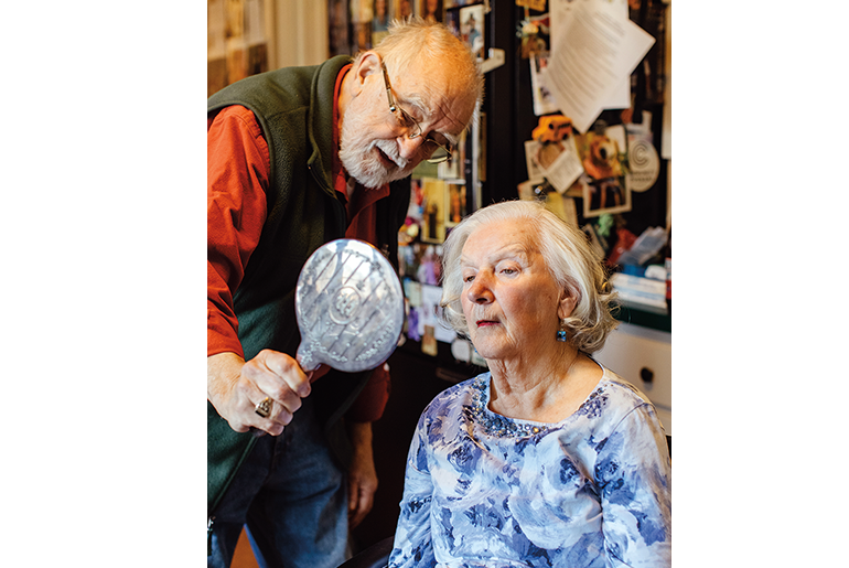 Image of a man with white hair, glasses and a beard in a dark grey vest and red shirt holding a hand mirror for a woman with white hair and a blue flowered shirt 