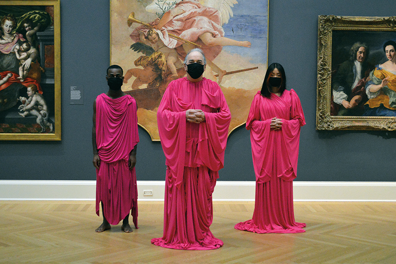 Image of three figures in pink robes and black masks in front of large format paintings in a museum setting 