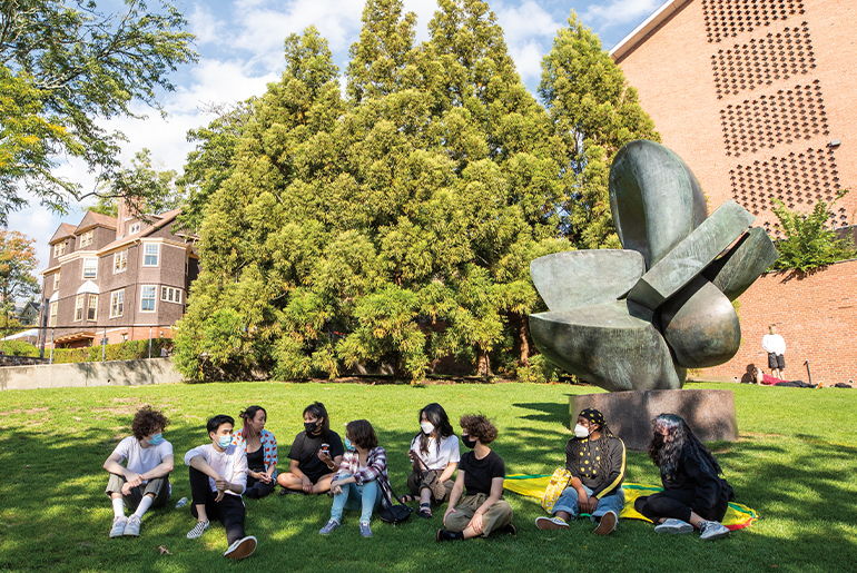 An image of RISD students, in masks, sitting on the grass outside of a building on a sunny day.