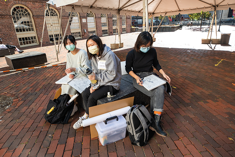 Students under a open tent with masks working on sketch pads