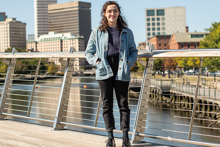 Image of a person in a jean jacket and black pants on a bridge over the Providence River