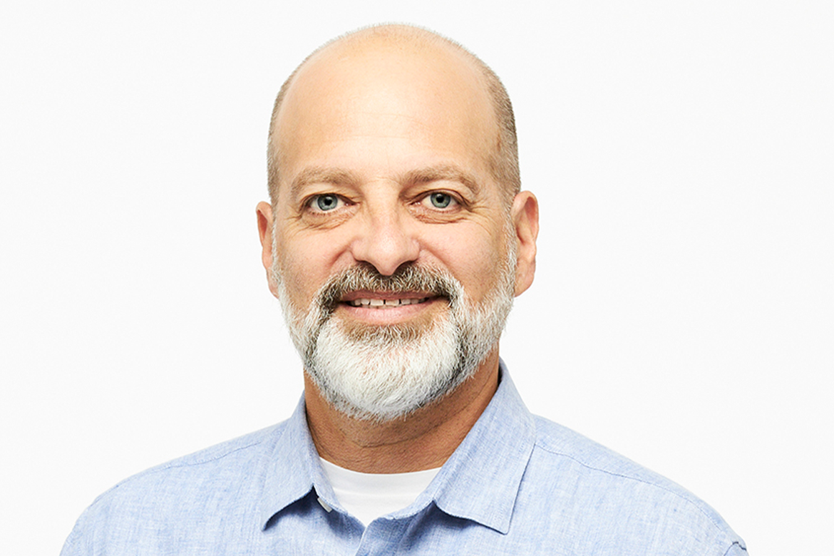 headshot of a smiling balding man with a grey beard in a blue button down and white t-shirt