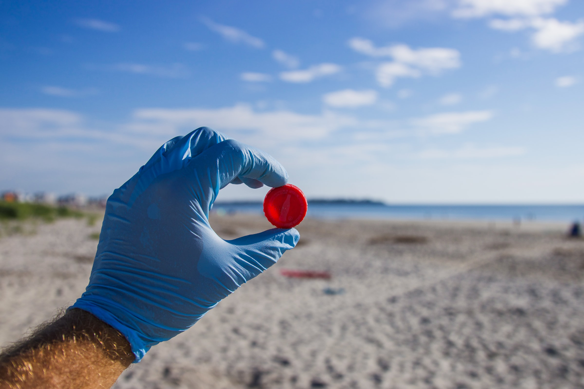 Image of a person wearing a blue nitrile glove holding a red bottle cap against a blue sky and beach sand