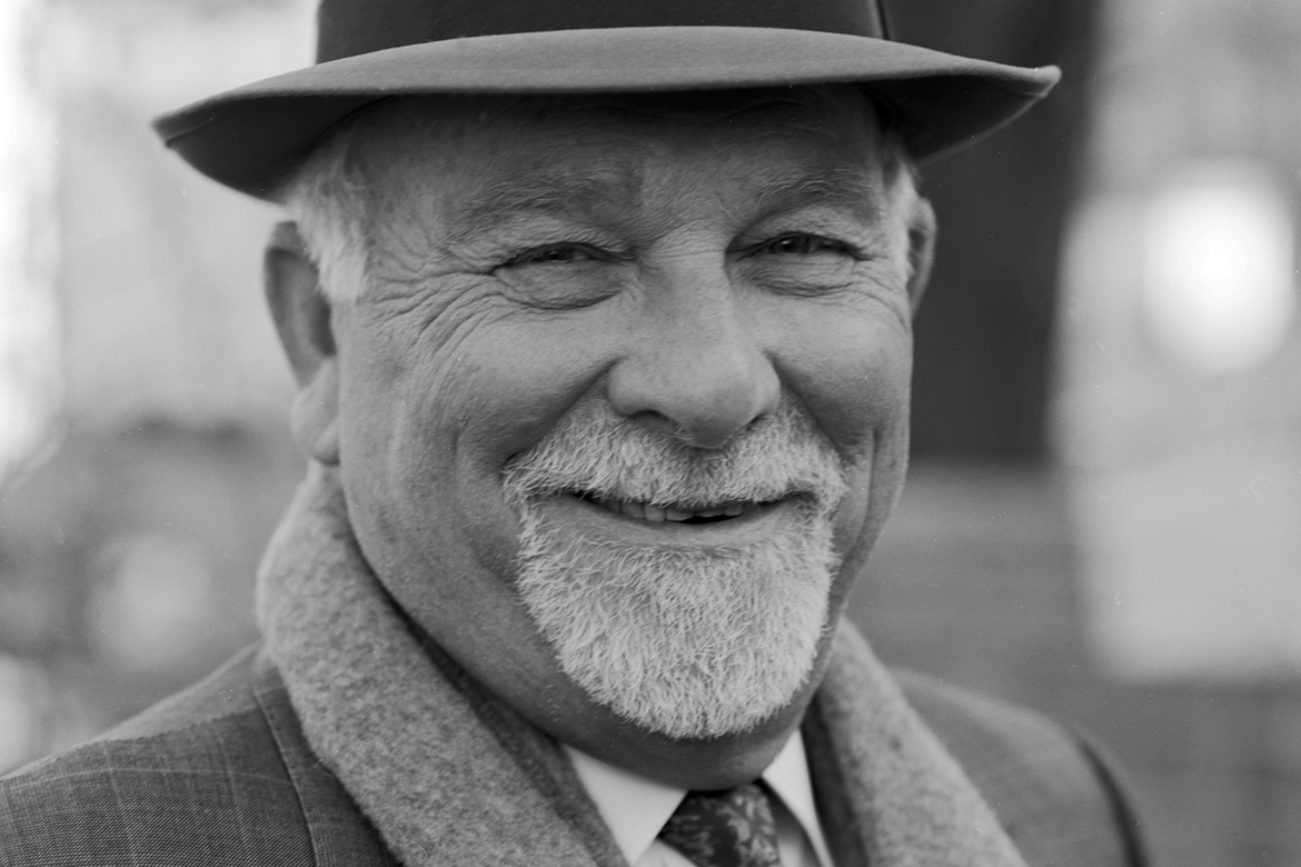 Black and white photo of a man with a white goatee fedora cap tie and scarf