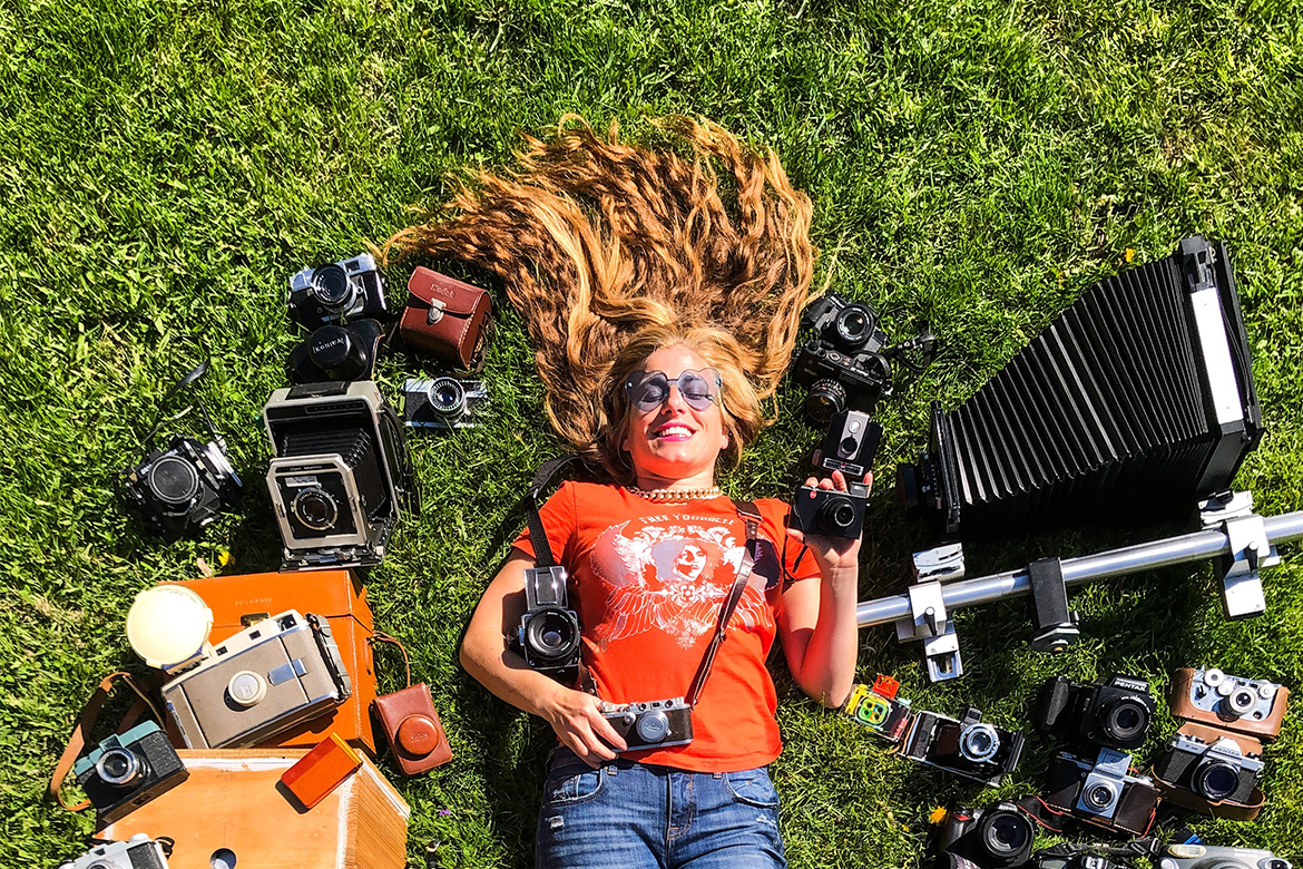 Jordanne Renner in an orange shirt and jeans, lying on a grass field and surrounded by cameras