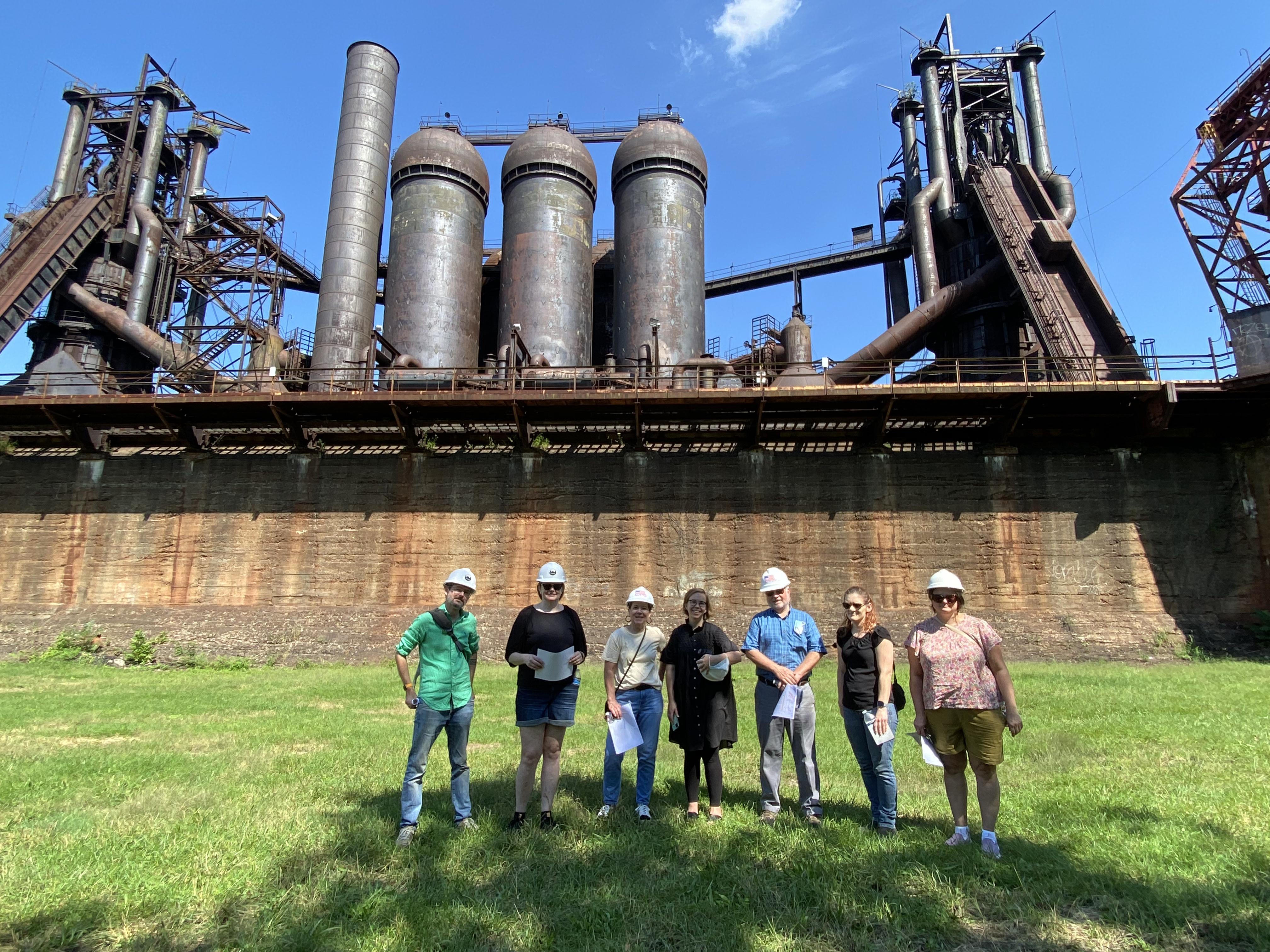 Six people in construction hats standing outdoors in front of a brown furnace
