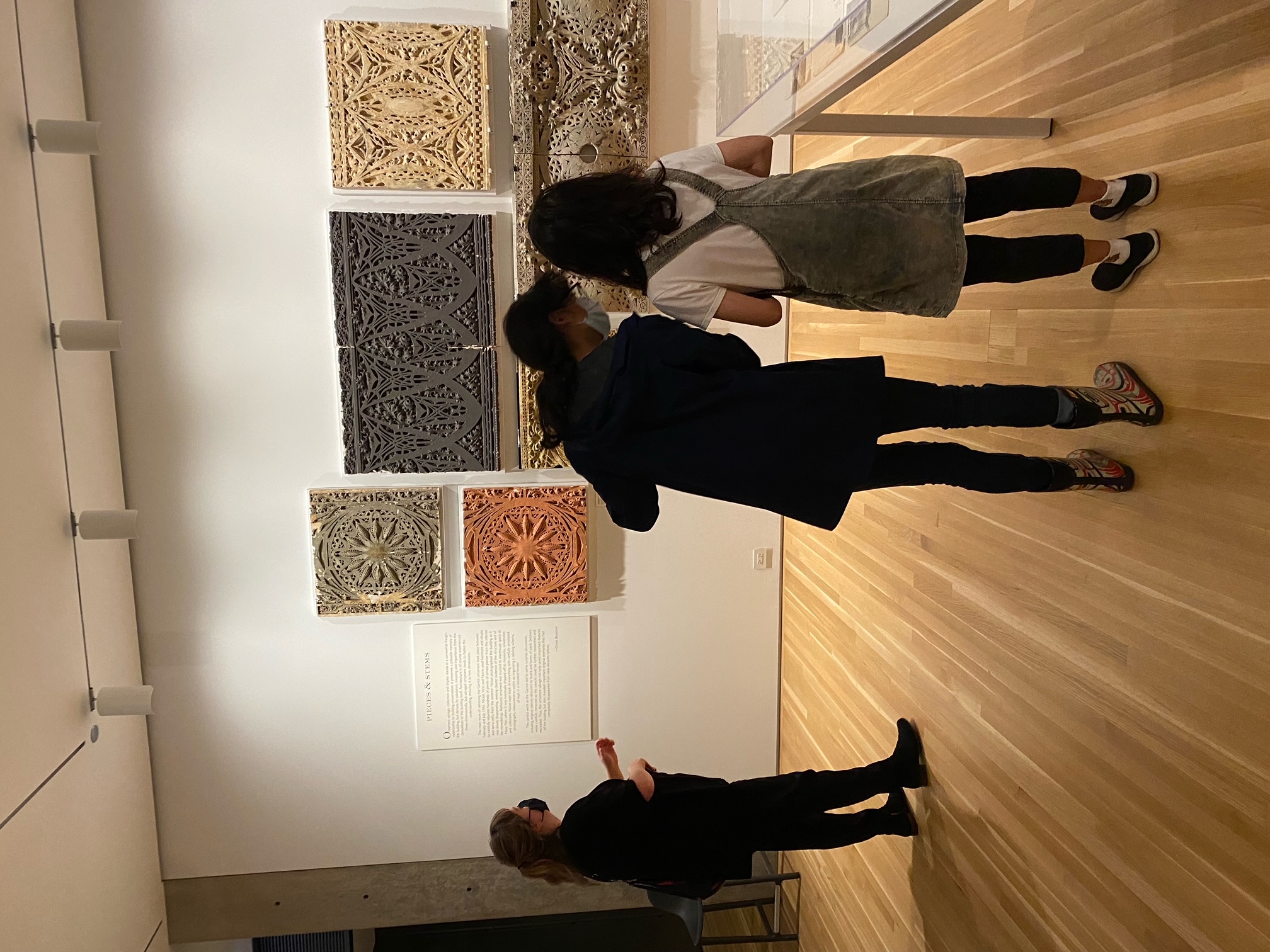 Three people examining art pieces on a white gallery wall that depict spiraling and floral designs
