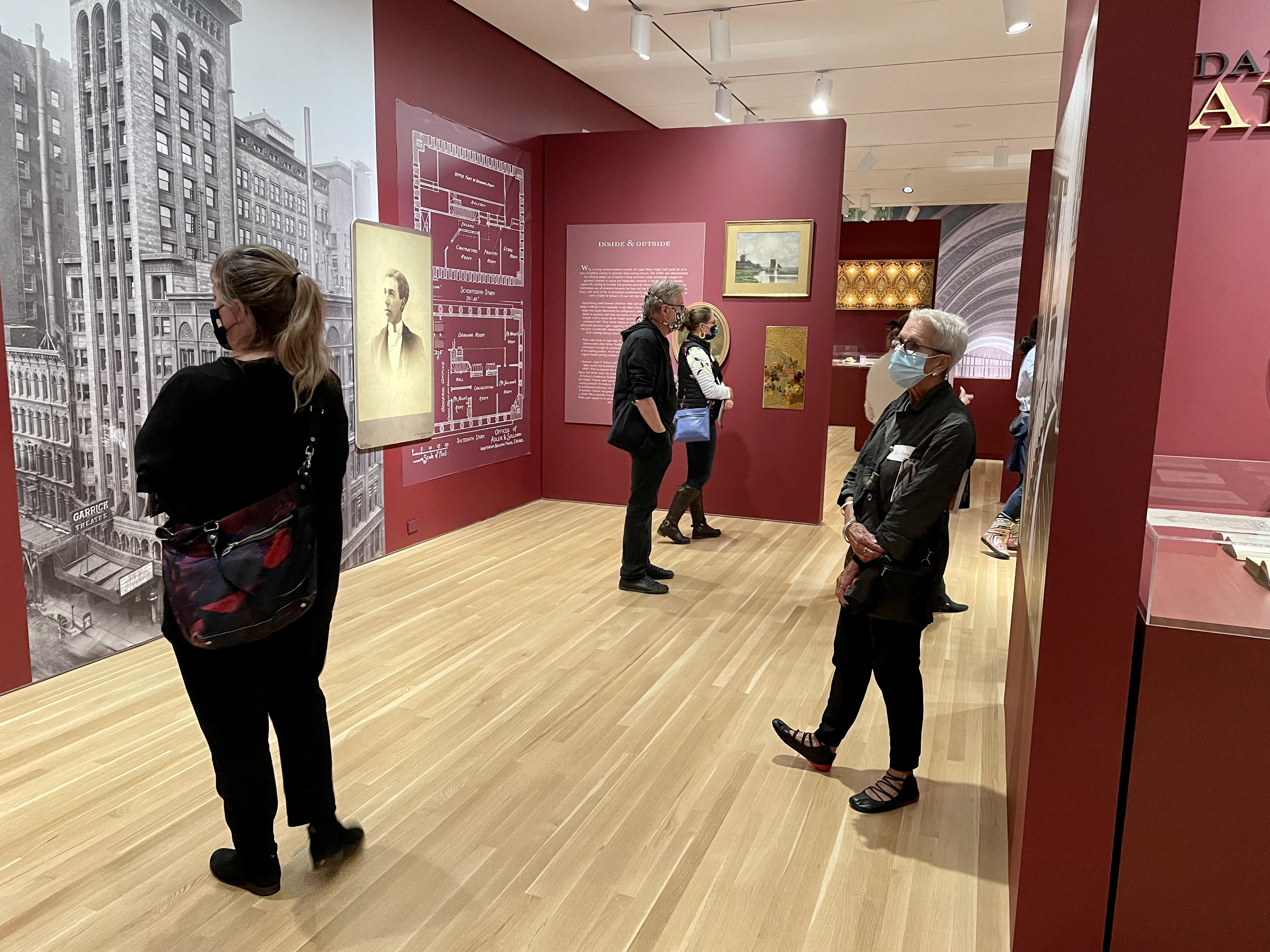 Six people standing in an art gallery with red walls and looking at black and white photographs of architecture