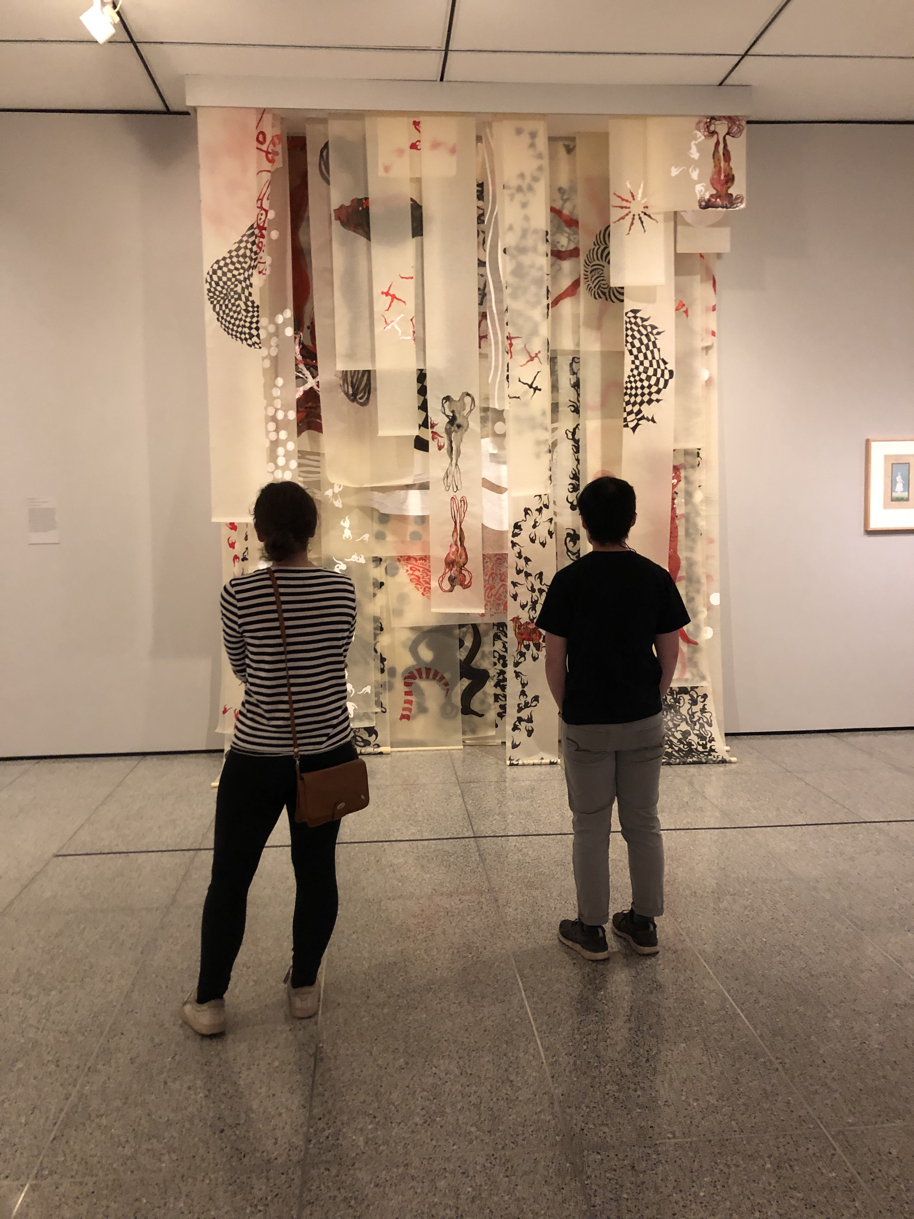 Two people examining an art piece made of white strips with black and red patterns and hanging from the ceiling