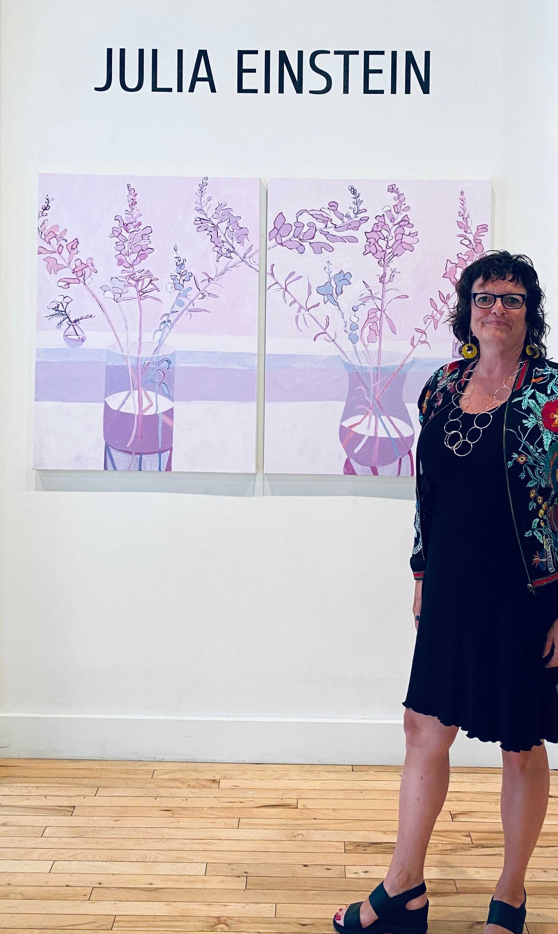A smiling standing in front of two lilac and lavender paintings of flowers