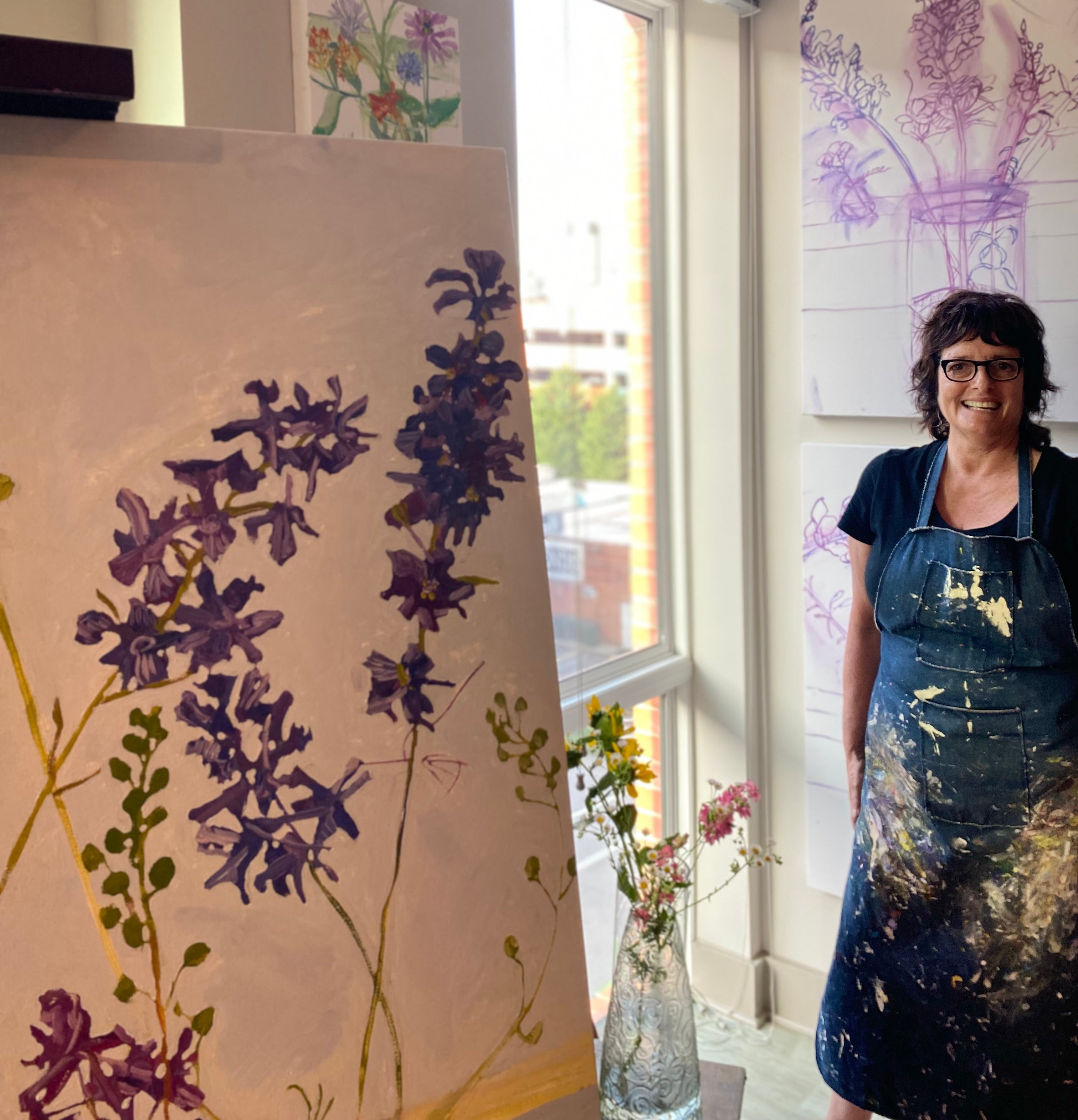 A smiling and standing person surrounded by paintings of purple flowers