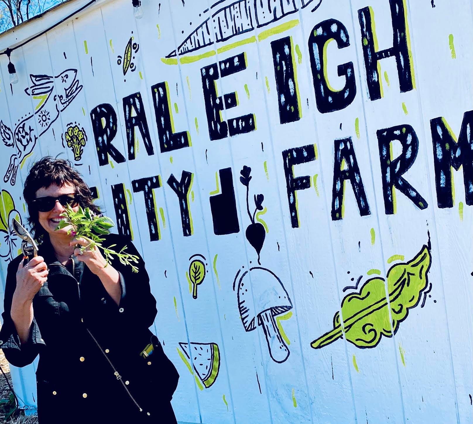 A smiling person holding shears in a plant in front of a Raleigh City Farm sign