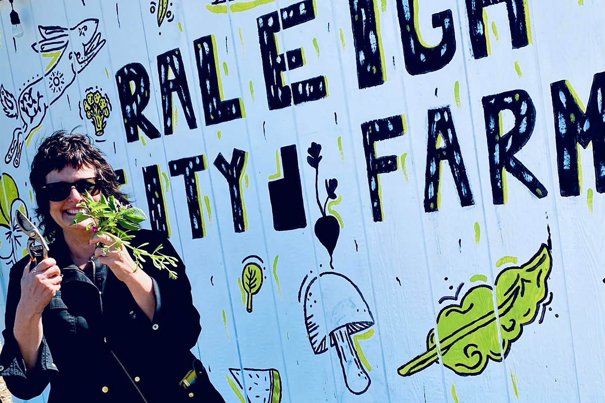 A smiling person holding shears and a plant in front of a Raleigh City Farm sign