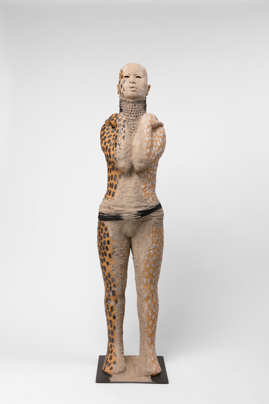 A tan life-sized sculpture of a person standing with their hands on their shoulders