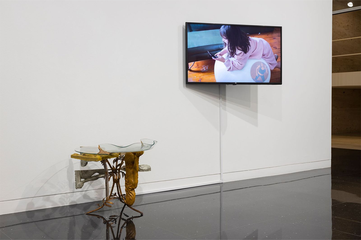 An exhibition hall holding a glass table with metallic elements and a television showing a woman on a cushion