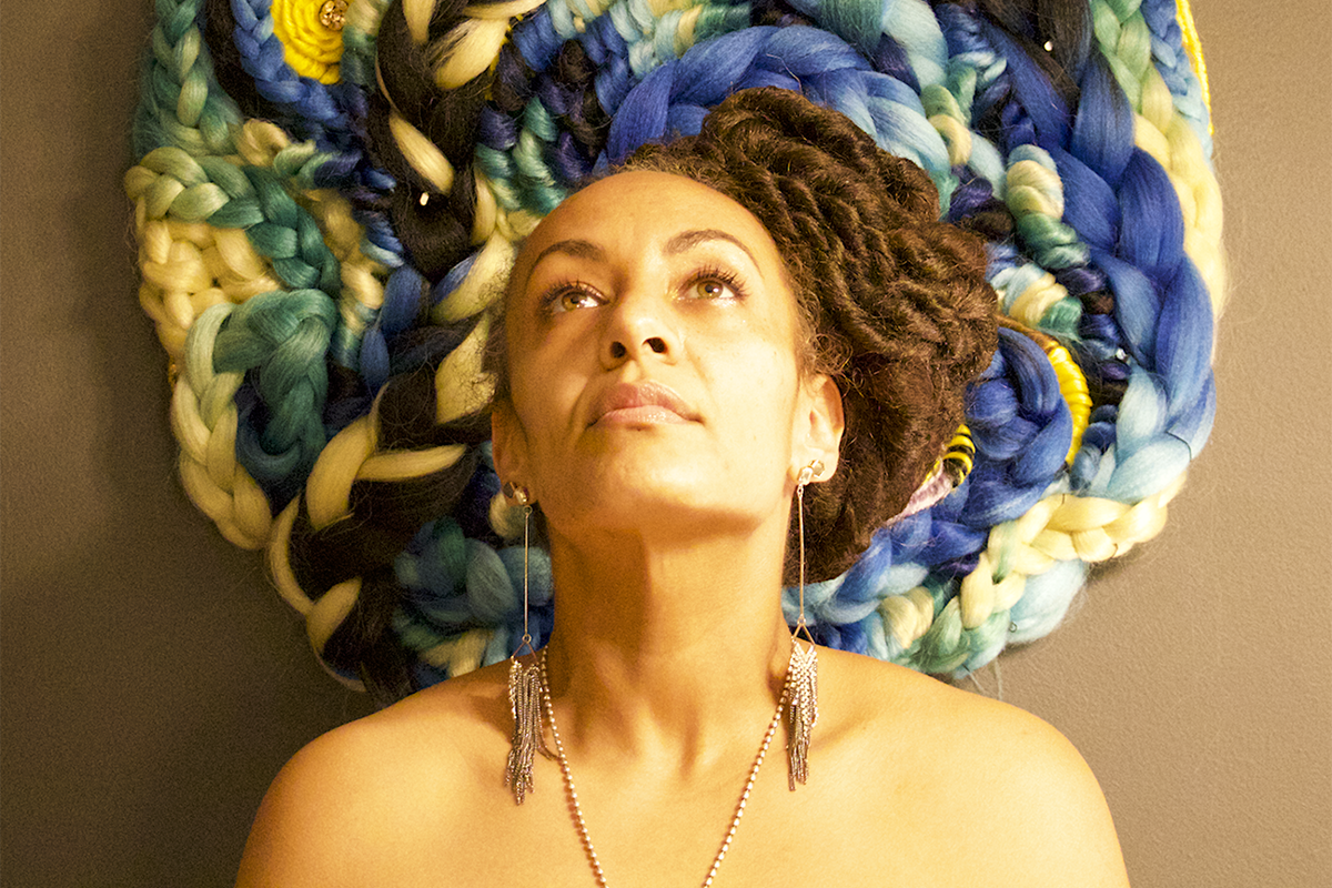 Image of a female looking upwards with a braided blue yellow and green tapestry behind her