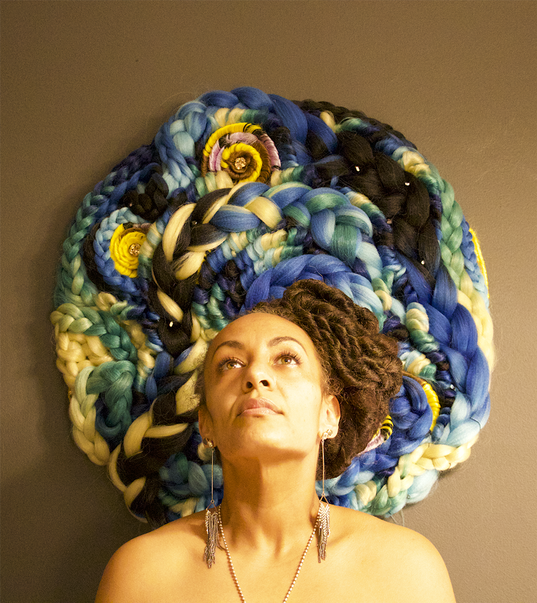 Image of a female looking upwards with a braided blue yellow and green tapestry behind her