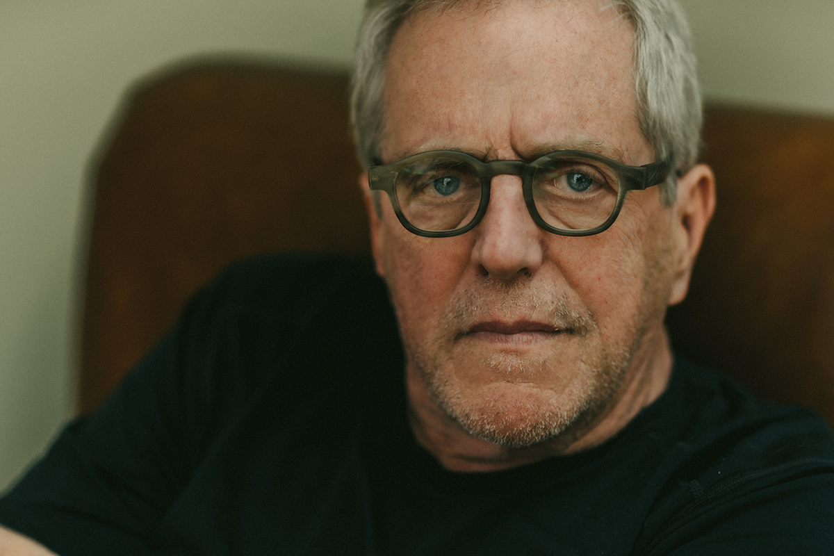 Image of an older man with grey hair and glasses sitting in an armchair with a dark sweater on