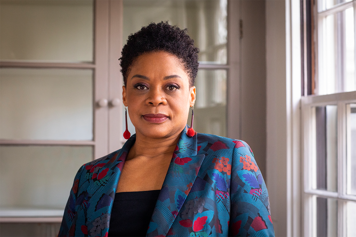 RISD President Crystal Williams in a bright blue multicolor blazer, red earrings standing in an office setting 