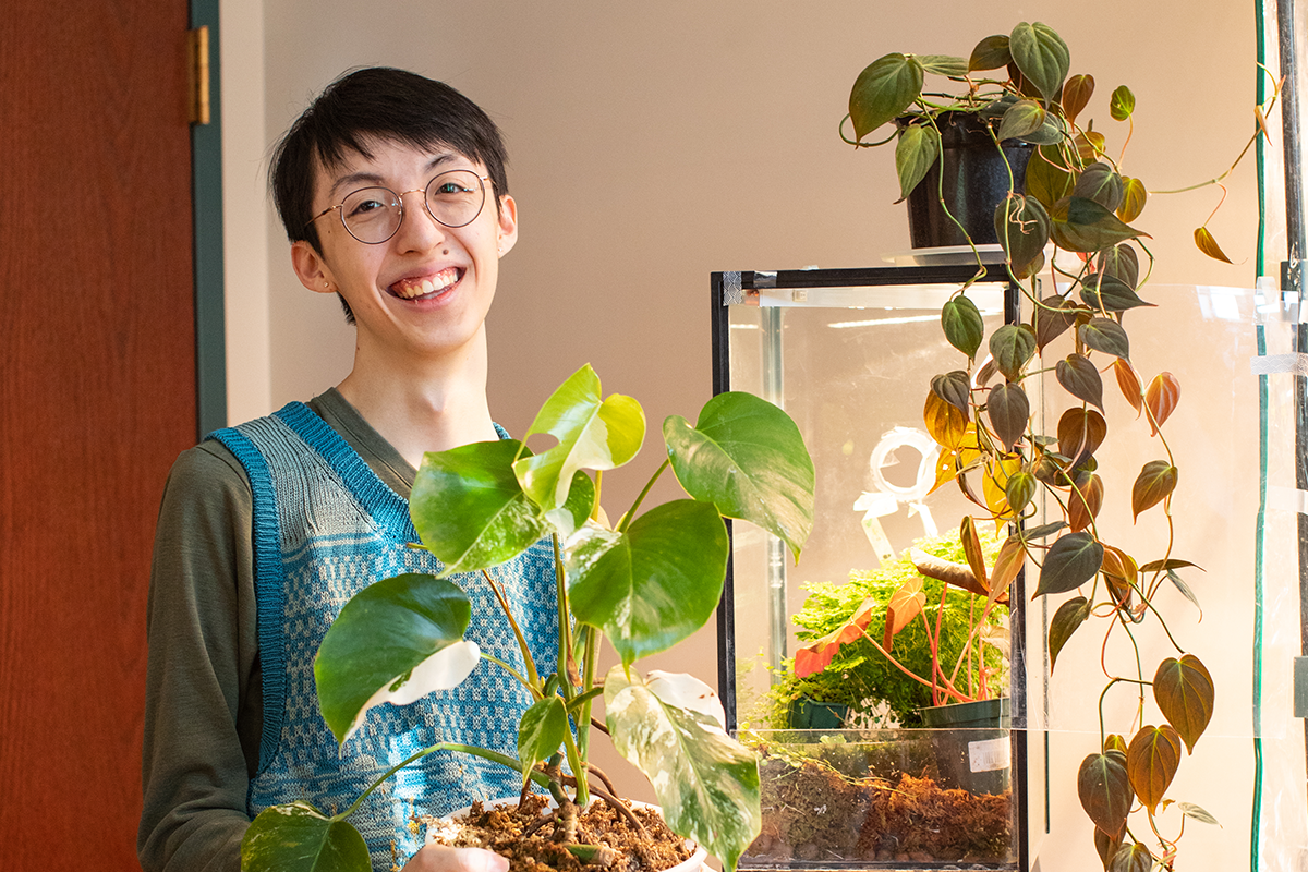 Image of a student with a blue shirt and round glasses standing with plants
