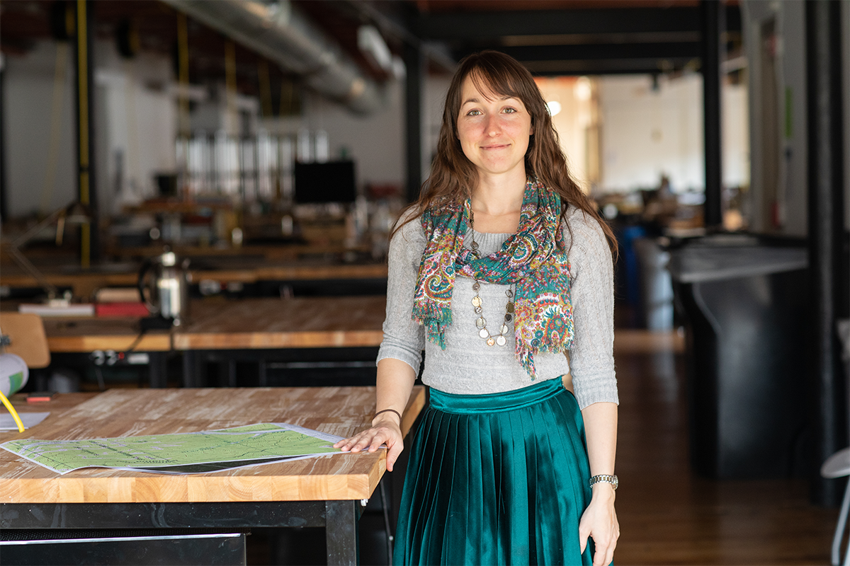 Female in a bright green pleated skirt with a grey sweater and multicolored scarf standing next to work benches in a studio setting