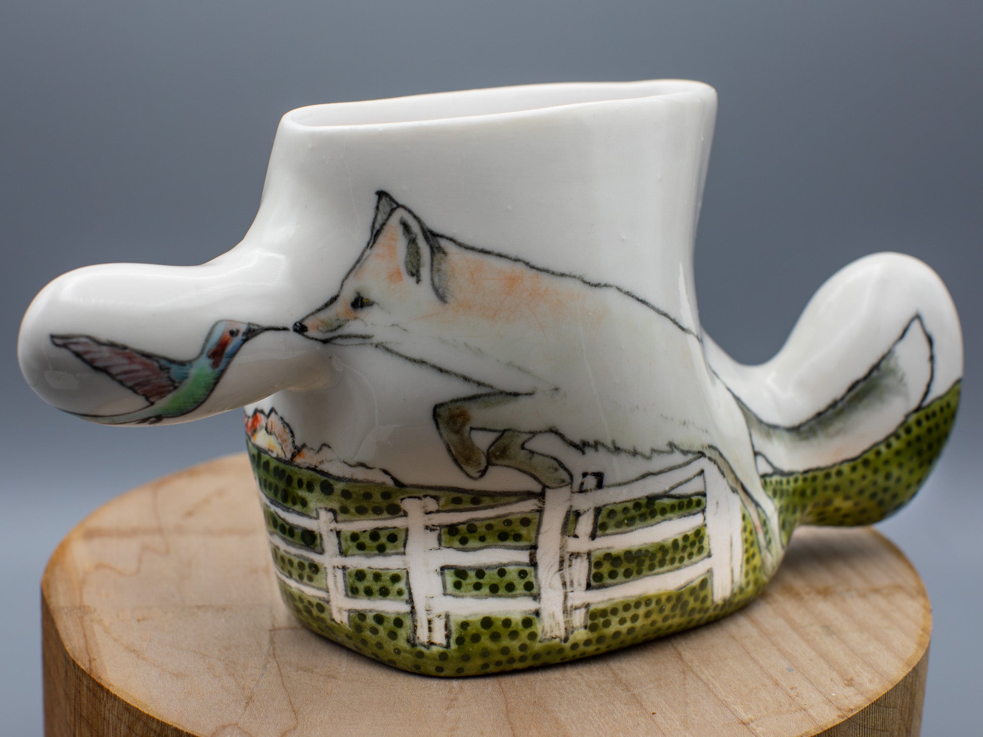 A vase with a painting on its side of a fox touching its nose to a hummingbird's beak