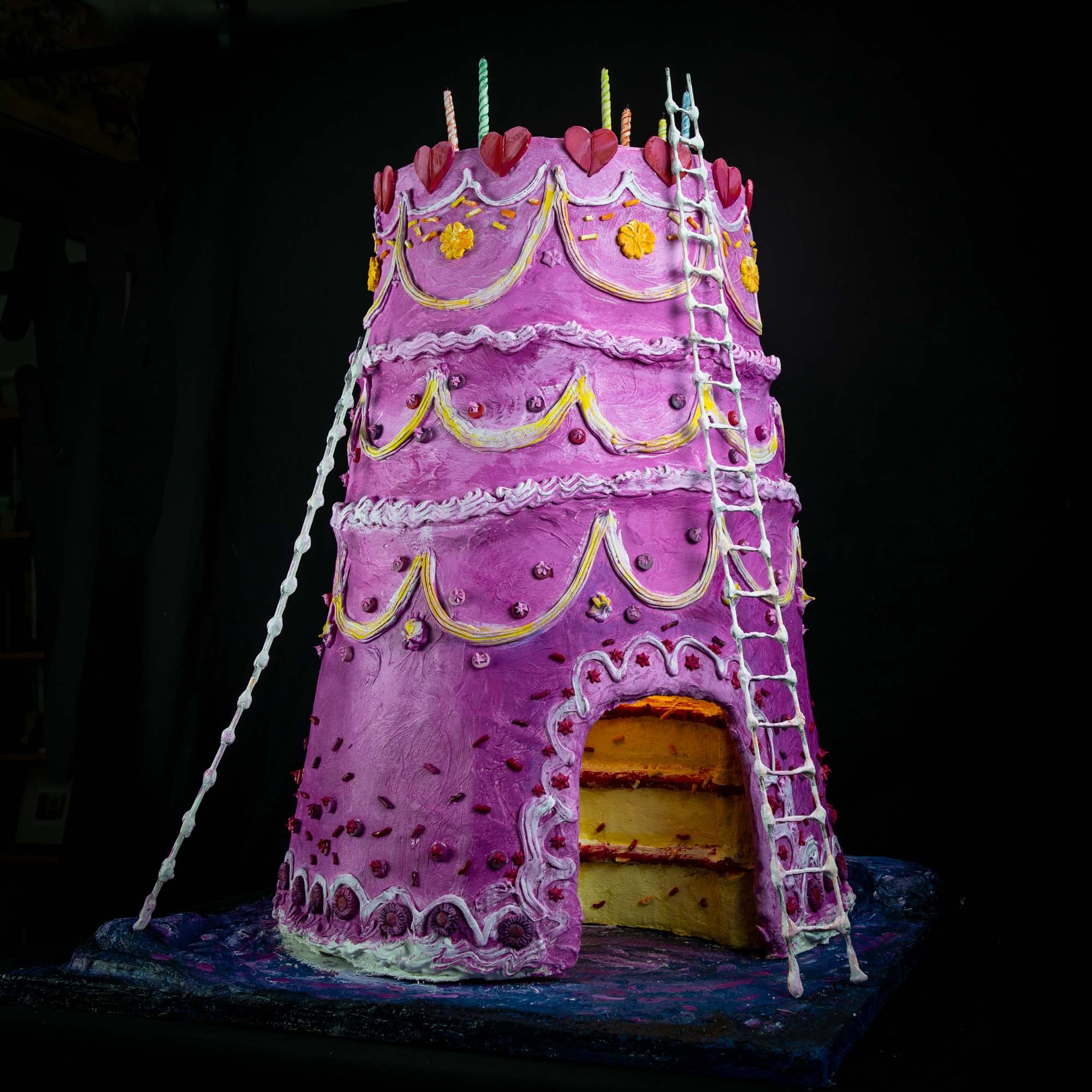 A diorama of a pink three-tier birthday cake with ladders resting against its sides