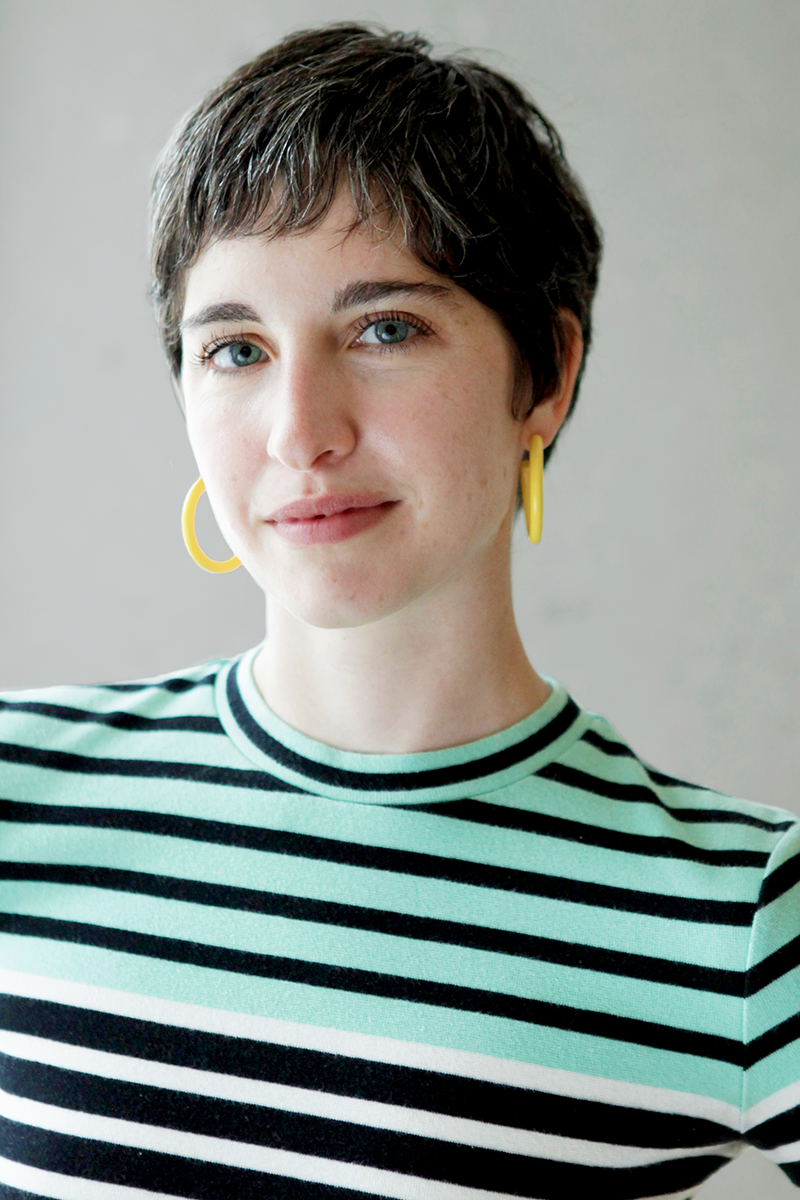 Emily Ann Hoffman wears a striped green and black shirt and yellow hoop earrings in front of a plain white backdrop. Emily has short dark hair and blue eyes.