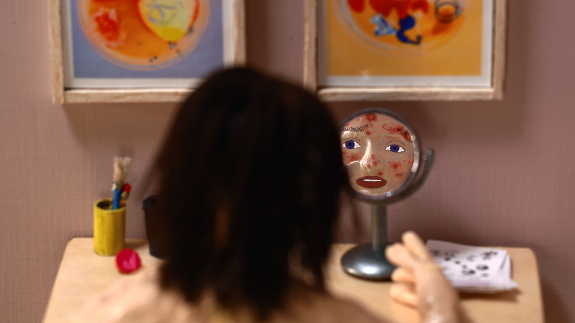 An individual observes their reflection and facial blemishes while sitting at a desk in front of a pink wall.