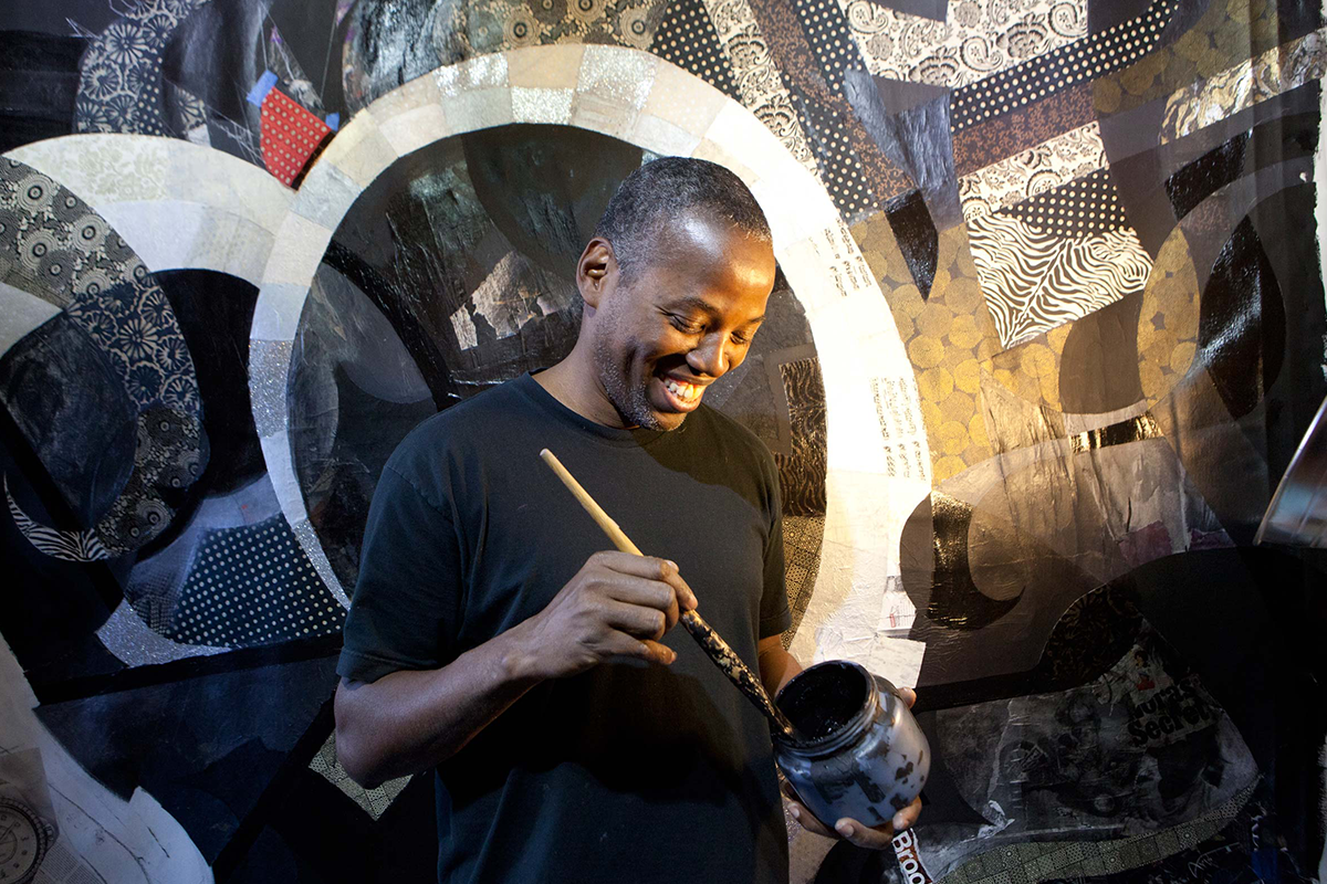 A smiling artists looking down at a paint container and brush in this hands as they are working on a mural with patchwork detail