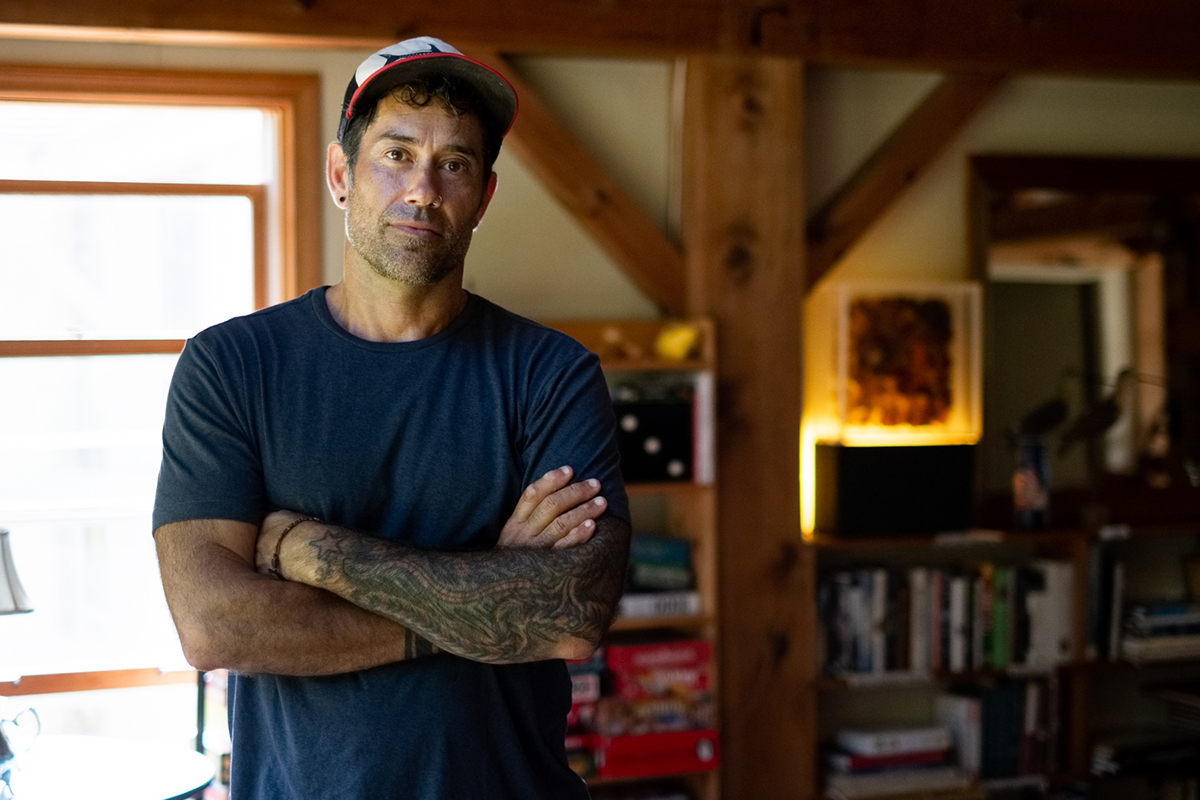 Jared Williams in a blue t-shirt standing with crossed arms in a country inn library