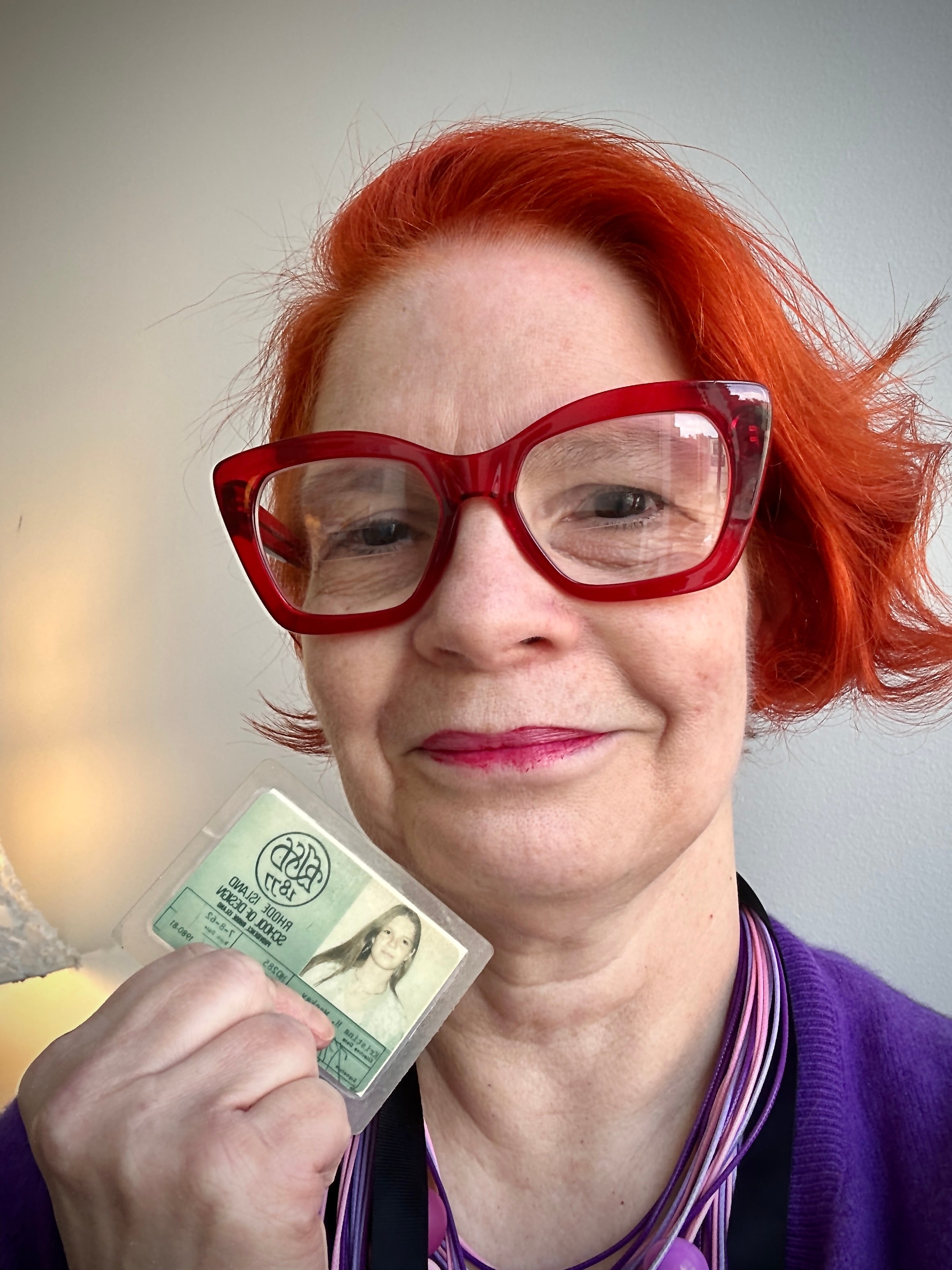 Kristina Lavoie with red hair and red framed glasses holds her RISD ID from her days as a student