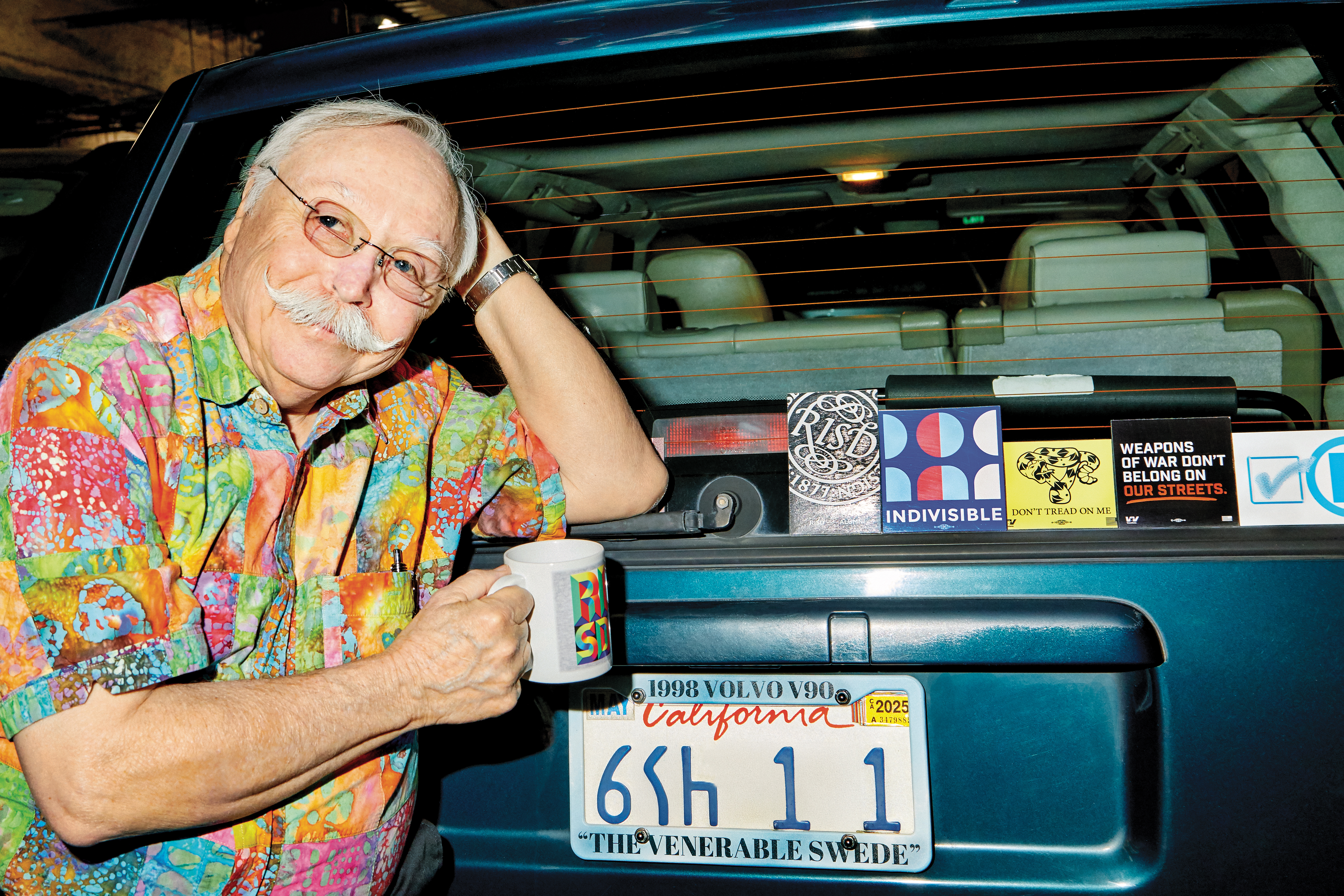 Bill Kienzel 67 ID stands behind a dark blue vintage station wagon is. a brightly patterned Hawaiian-style shirt holding a coffee mug.